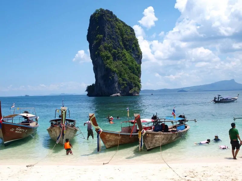 Boats line up along a beach on Koh Poda while people swim around them in Thailand.
