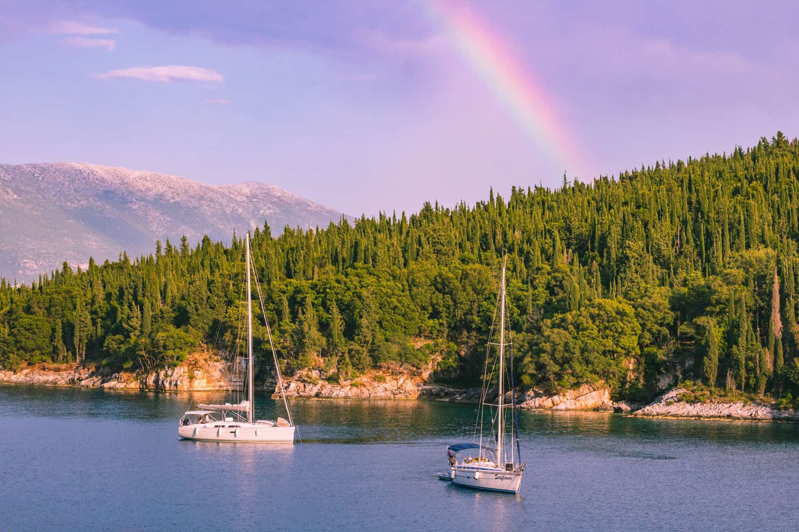 Two sailing boats anchored just off shore with a rainbow behind them in the sky in Greece.