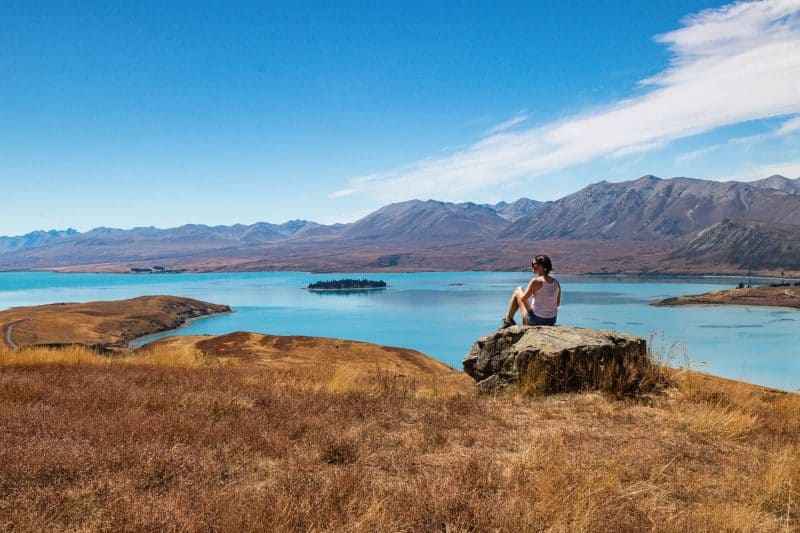 Mount John Trail, with good views of the blue lake and a woman on the rock - one of the best things to do in Lake Tekapo