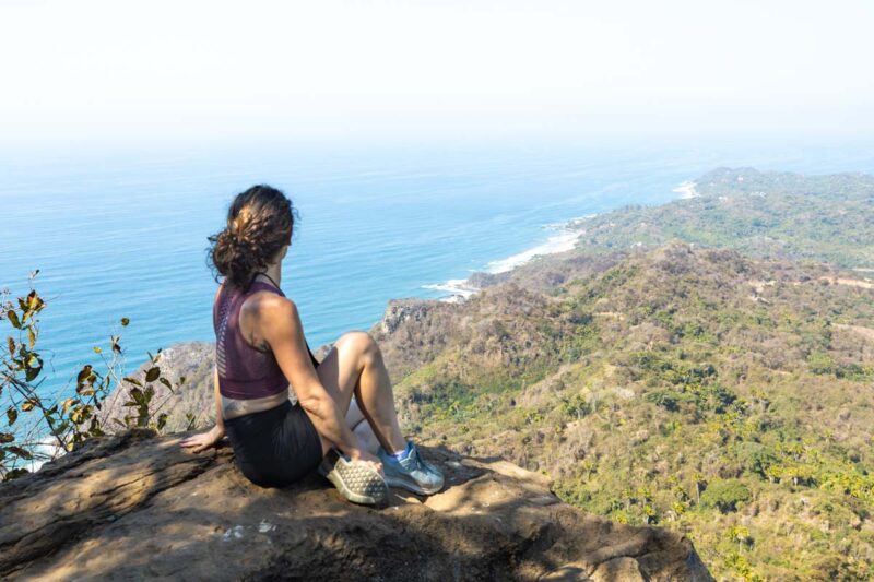 Nina sitting on rock on top of Monkey Mountain and looking out over ocean and coastline - hiking Monkey Mountain is one of the things to do in Sayulita