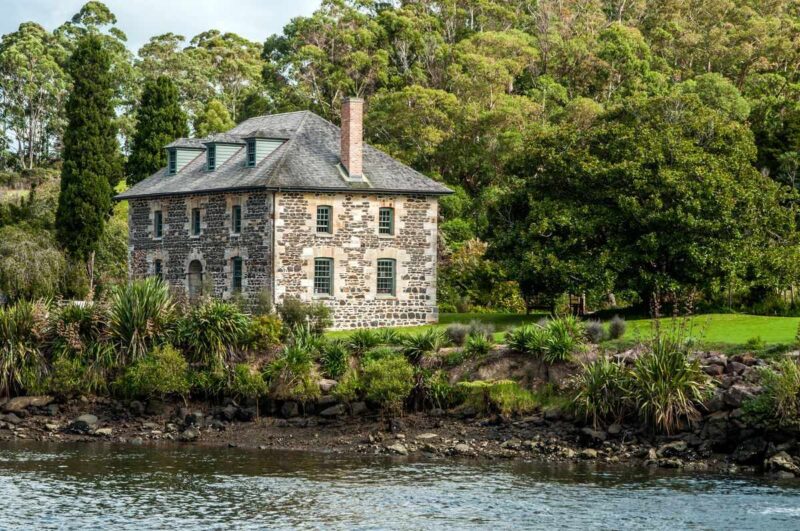 Old stone two story house on river bank with trees around it in Kerkeri, Northland