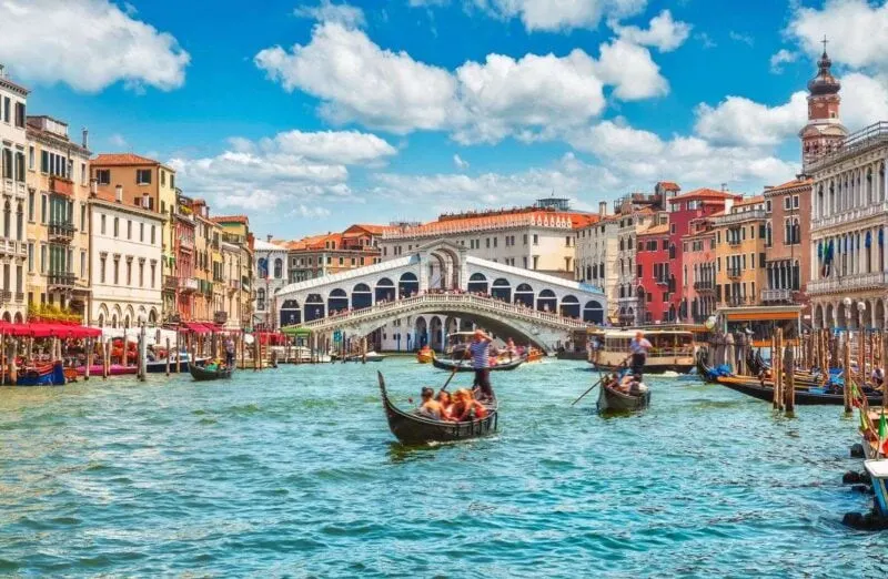 Rialto Bridge over Grand Canal with gondolas on canal and historic buildings on each side - add Rialto Bridge to your Venice Itinerary