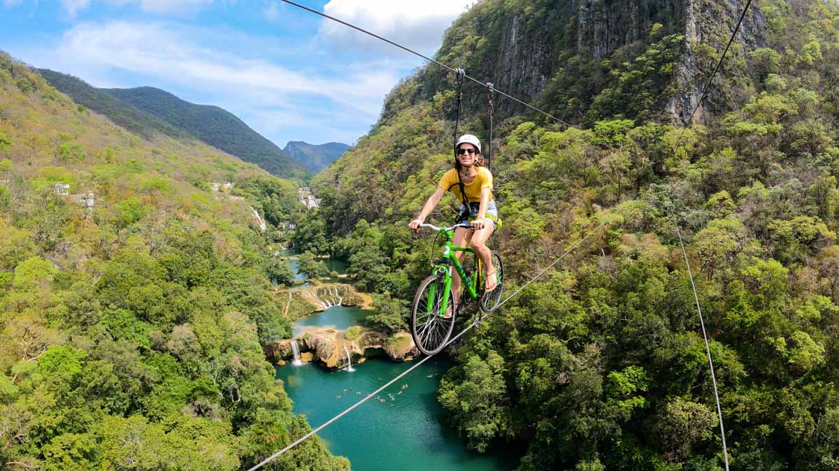 Nina riding a green bike across a zipline with views of Micos waterfall and jungled mountains in background.