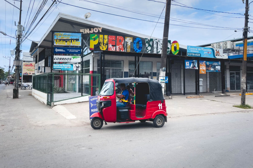 A bright red tuktuk driving in front of a huge "Puerto Viejo" sign.