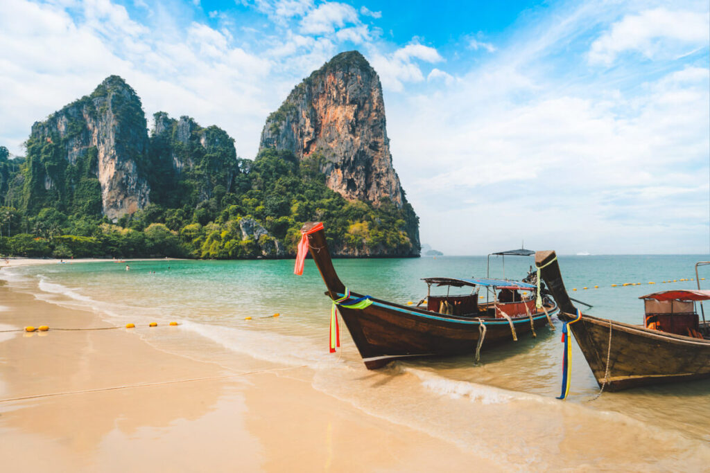 A couple of fishing boats dwarfed by huge limestone rocks in the background and bright blue ocean waters of Railay West Beach near Krabi.