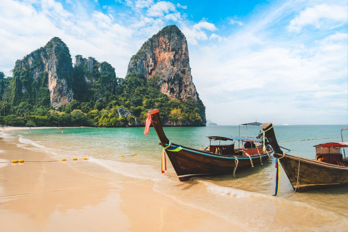 A couple of fishing boats dwarfed by huge limestone rocks in the background and bright blue ocean waters of Railay West Beach near Krabi. A couple of fishing boats dwarfed by huge limestone rocks in the background and bright blue ocean waters of Railay West Beach near Krabi.