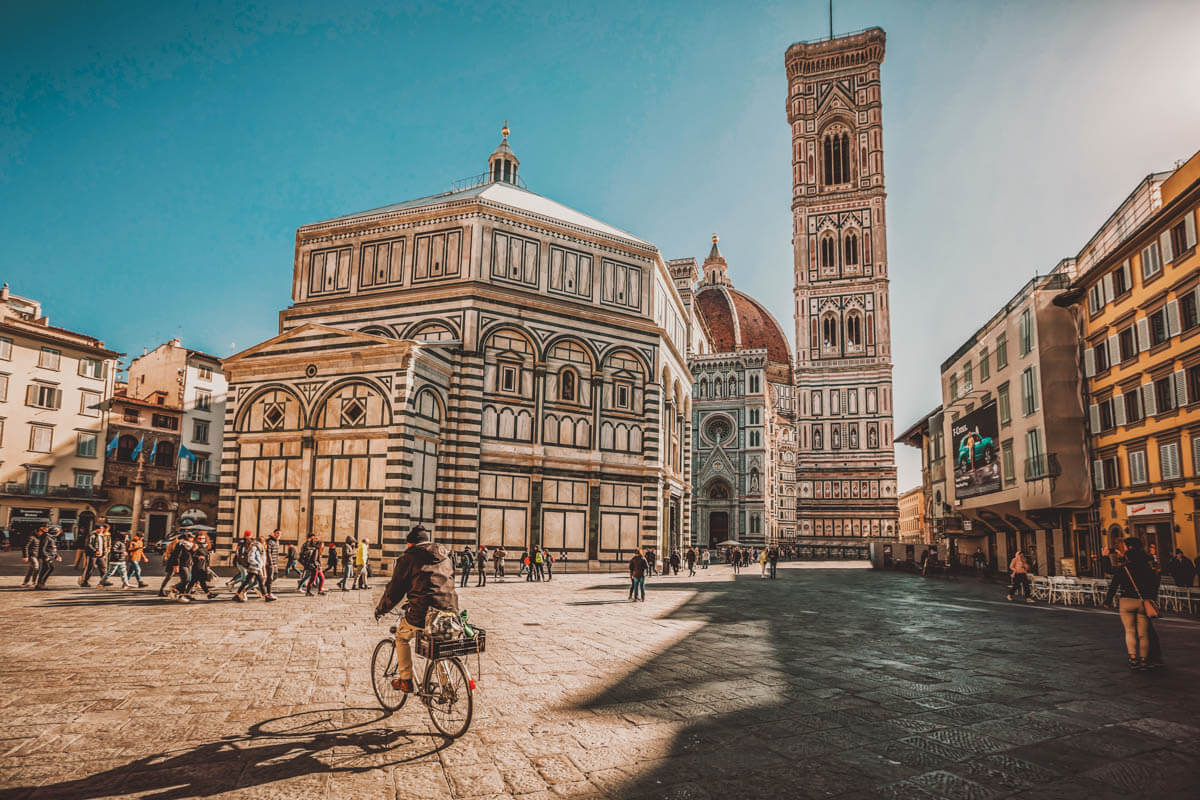 A plan rides a bike through the stunning Piazza del Duomo in Florence, surrounded by traditional Italian buildings.