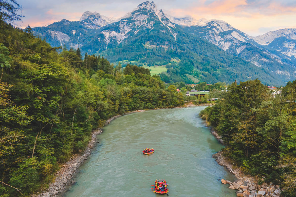 Two groups of river rafters on a wide river surrounded by trees in front of a backdrop of the Austrian Alps.
