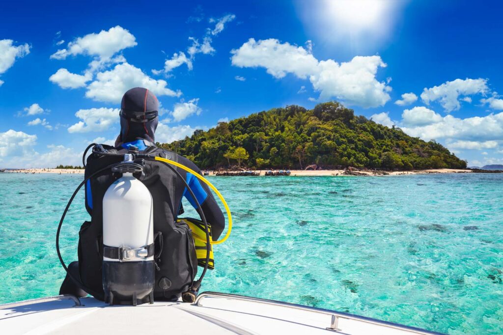 Man in scuba gear sitting on the edge of a boat besides a tropical island.