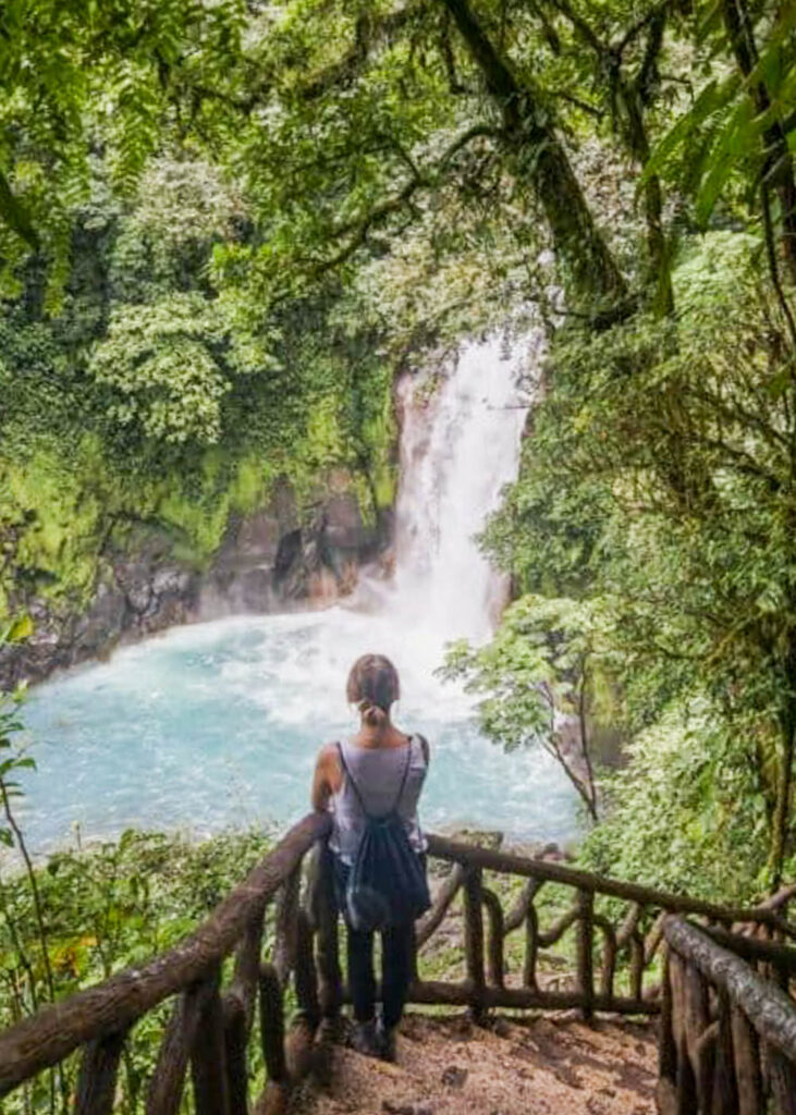 Woman standing on stairs overlooking Rio Celeste Waterfall.