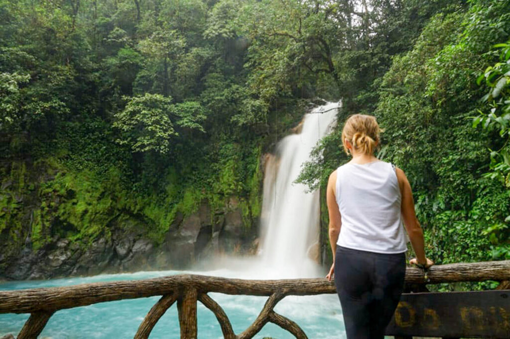 Woman leaning against wooden fence at Rio Celeste Waterfall.