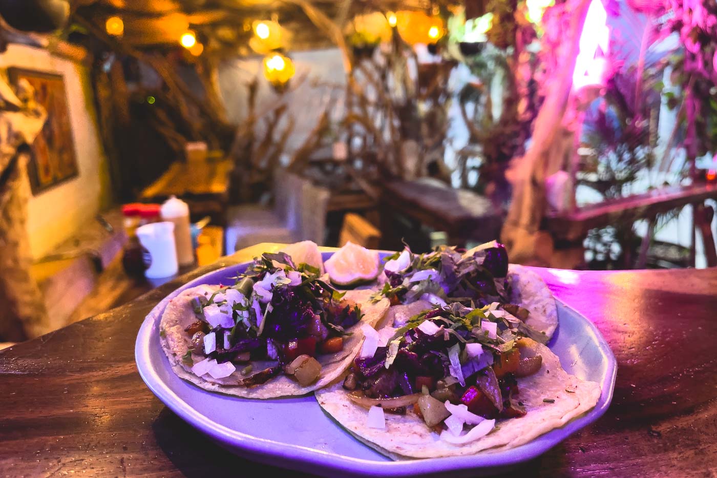 A set of 3 Chorizo tacos with a background of the funky lit restaurant called La Fogonera. 