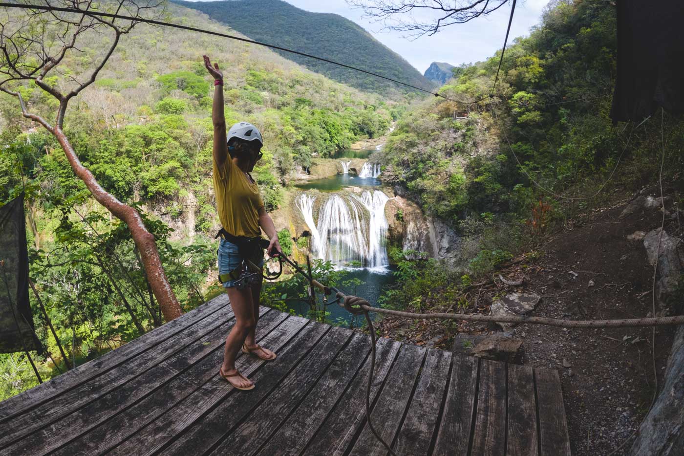 Nina with one arm raised in zipline gear and a helmet looking over Micos Waterfall from a wooden platform.