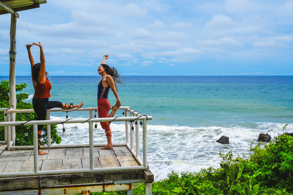 Two women stretching before a yoga session while standing on a wooden platform overlooking the ocean.