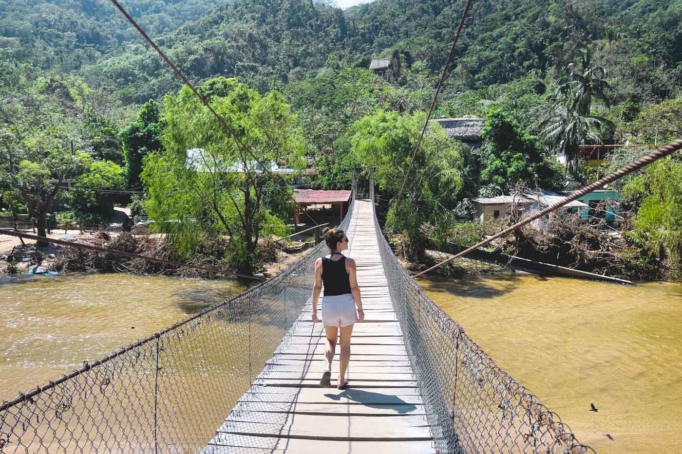 Nina crossing a long, wooden suspension bridge over a brown river heading towards the forests of Yelapa.