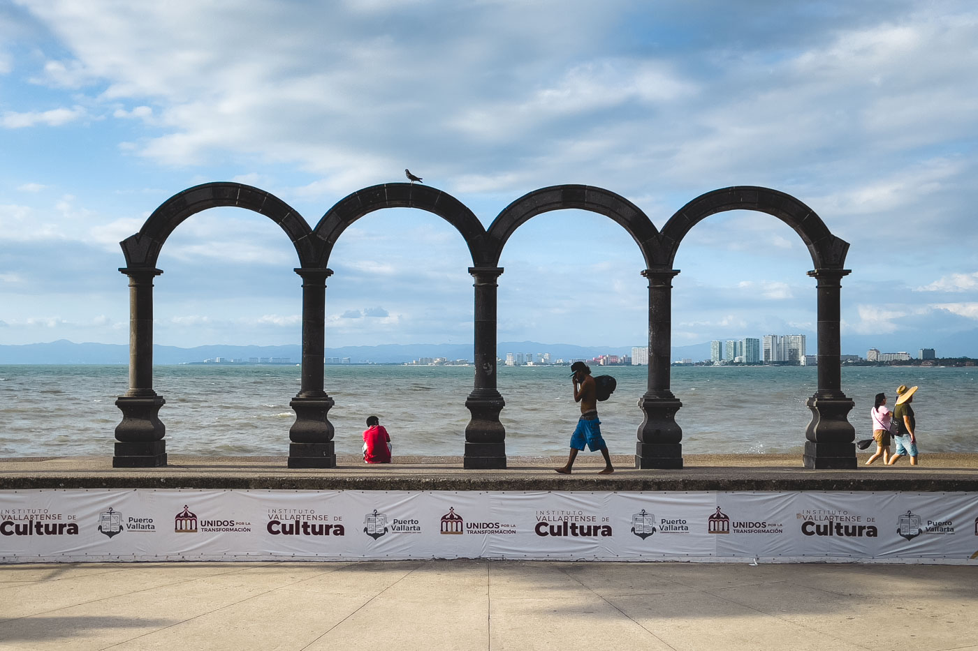 People walking past the Los Arcos Monument beside the ocean with a banner in front of it and Puerto Vallarta in the distance.