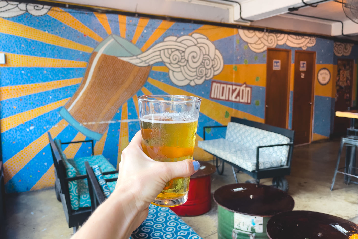 Nina's hand holding a pint of craft beer in front of a giant beer mural inside Monzon Brewery.