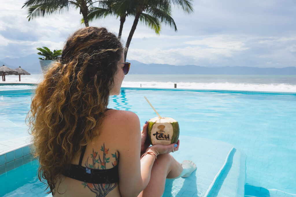 Nina sitting side a pool which overlooks the ocean while holding a coconut which reads "Tau Beach Club" on an overcast day.