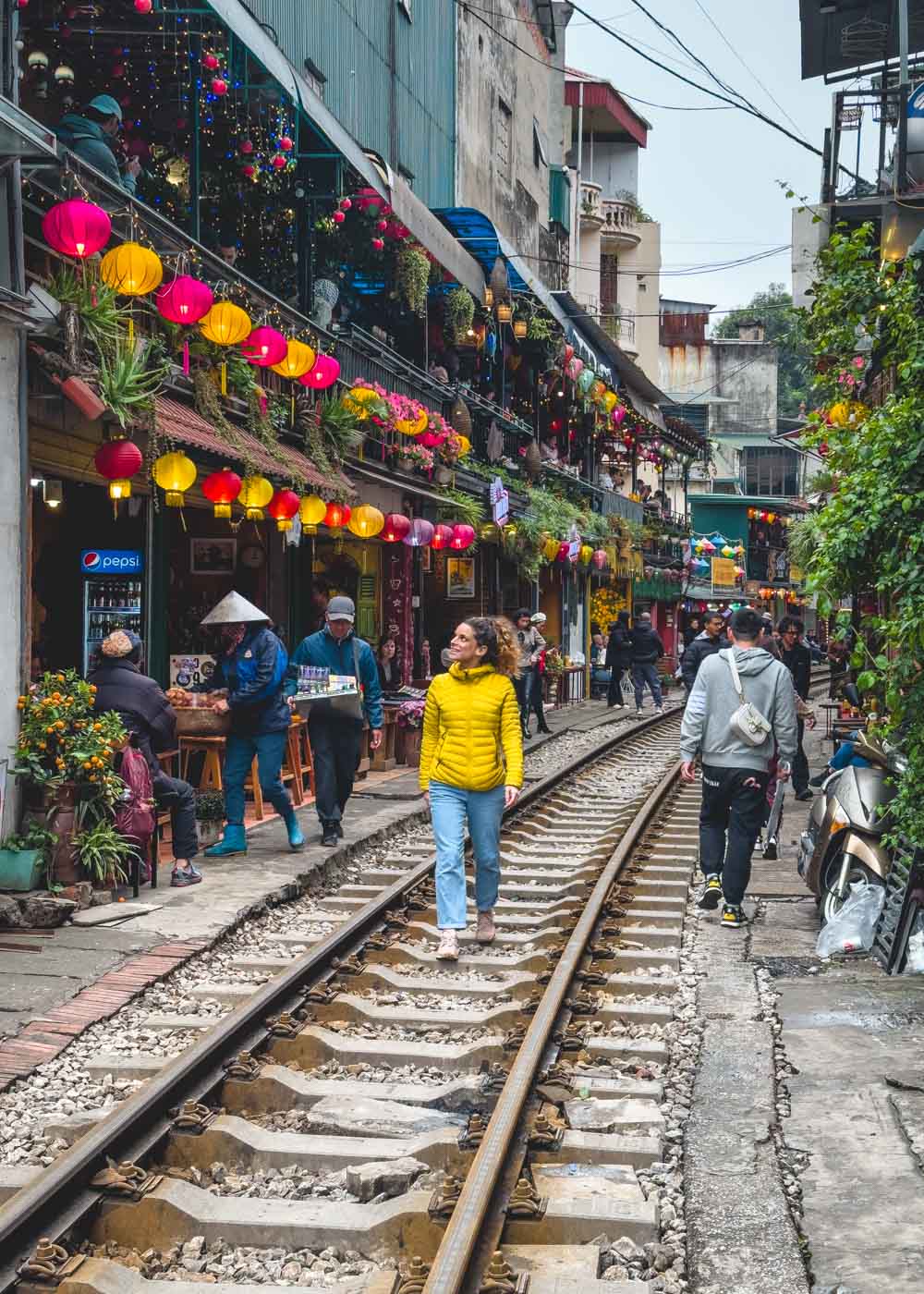 Nina in a yellow puffer jackets walking down the railway of Hanoi Train Street between cafes and lanterns.