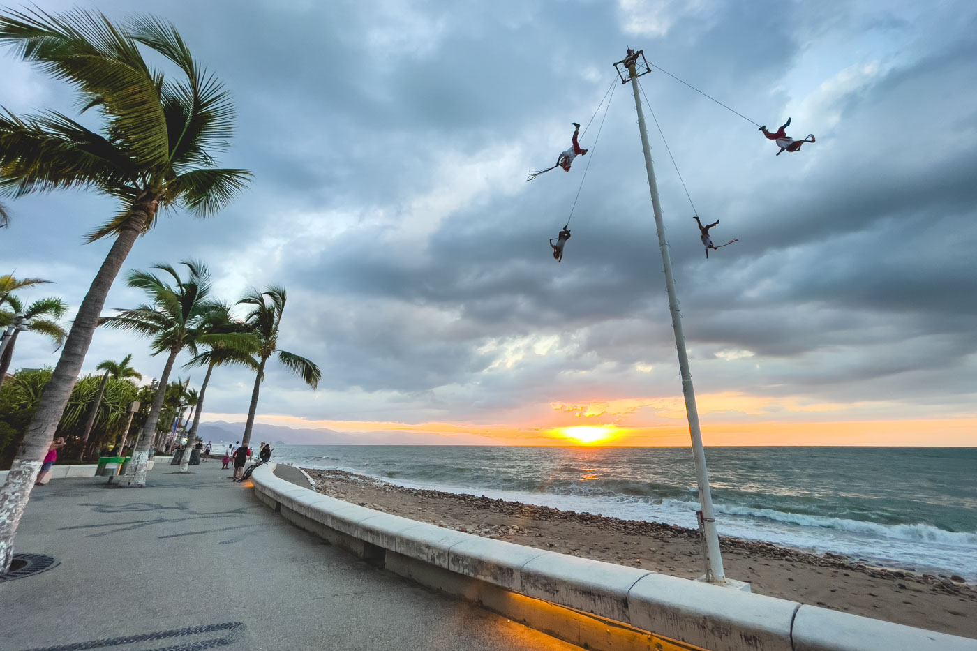 Four people suspended from a tall white pole by rope while swinging around above a beach at sunset.