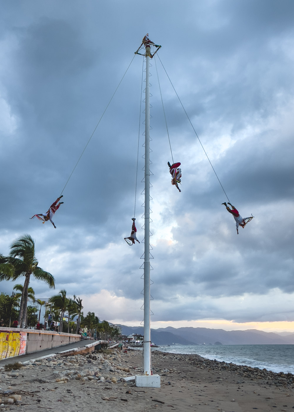 Four Papantla Flyers attached to a giant white pole on a beach.