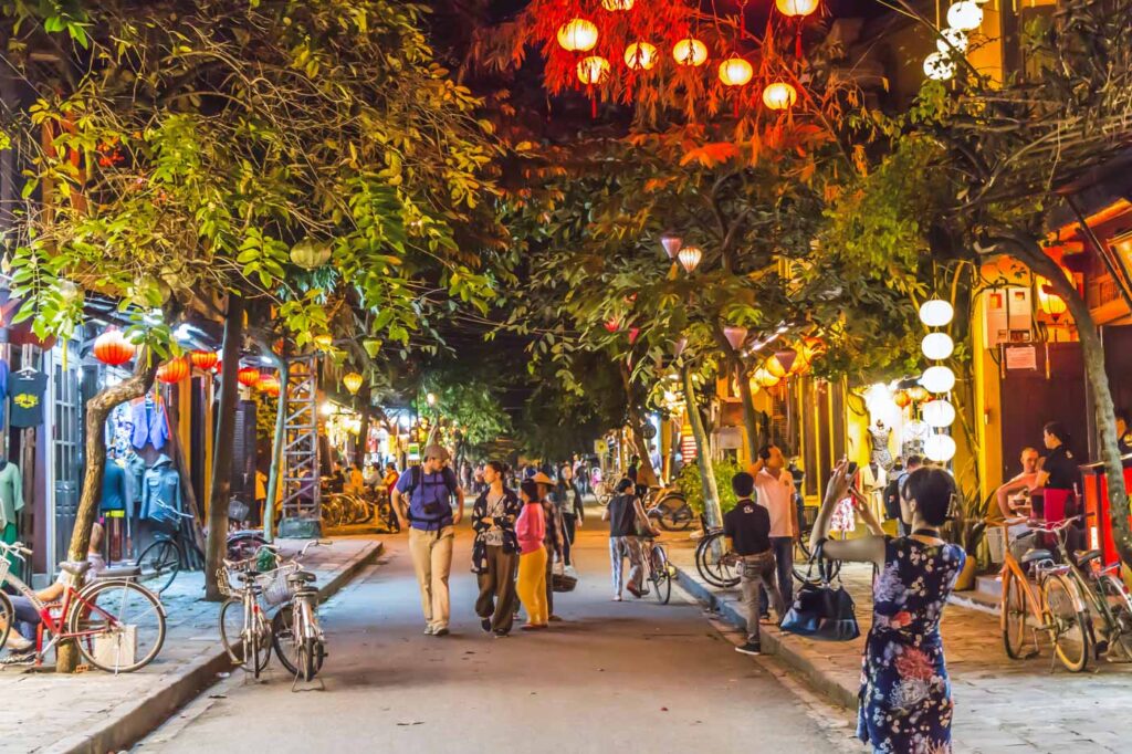Tourists taking pictures and admiring a street in Hoi An at night time under trees and lanterns.