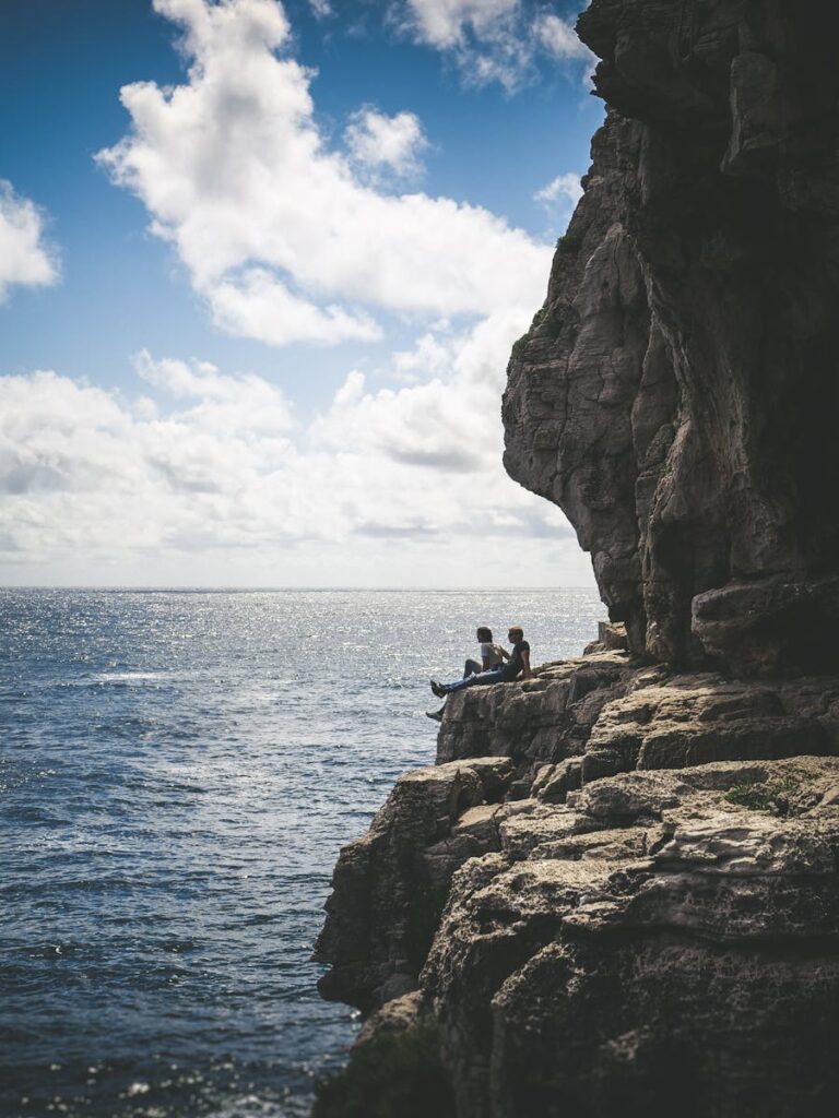 Couple sitting on a rocky cliff with scenic ocean view under a cloudy sky.