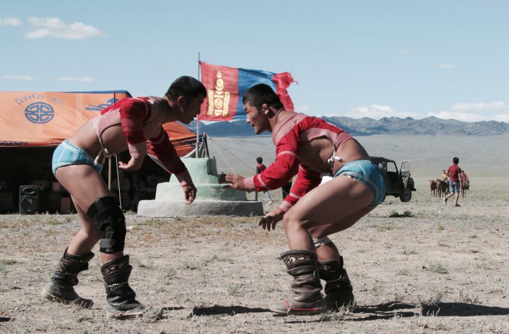 Two men compete in traditional Mongolian wrestling at a festival outdoors with cultural elements visible.