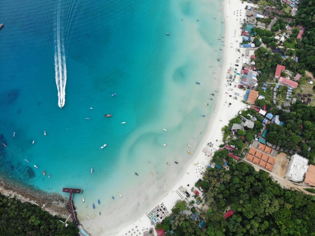 Drone view of shore with green forest and residential houses located in tropical island near sea with various boats on summer day