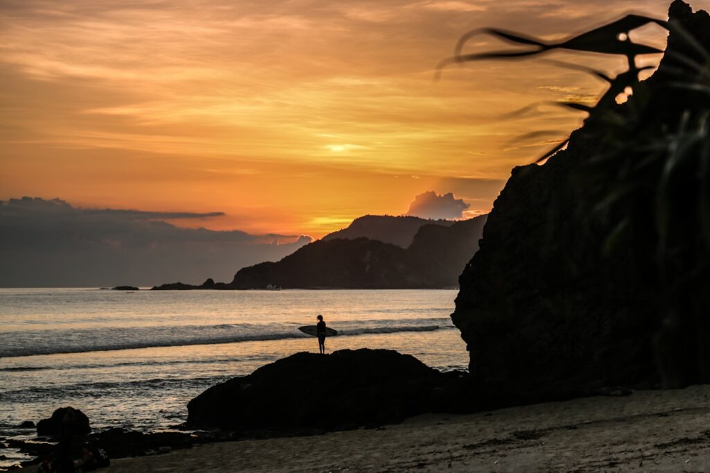 silhouette of person standing on rock near body of water during sunset