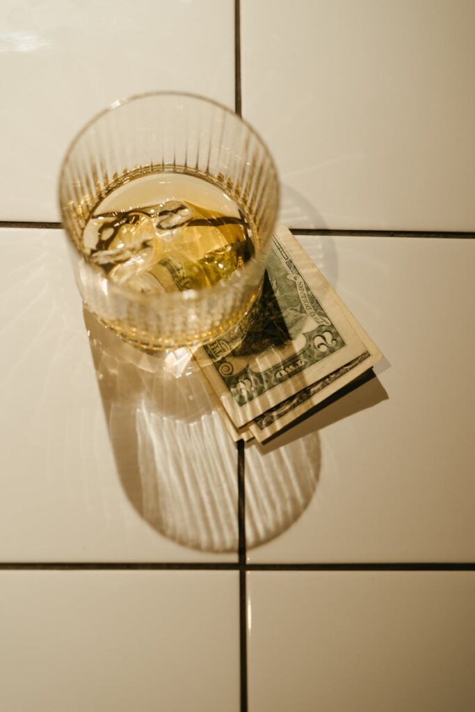A close-up of a glass of whiskey with ice on top of dollar bills, casting shadows on white tiles.