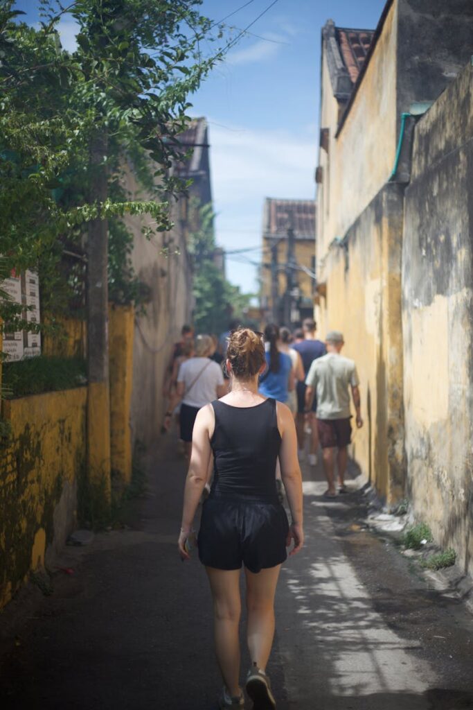 A group of people walk down a narrow alley between yellow buildings on a sunny day.
