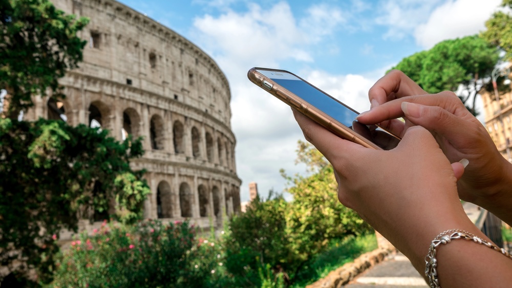 Woman messaging on the smartphone with the Coliseum of Rome at background. Girl having mobile phone conversation at city street. People travelers using device phone during travel
