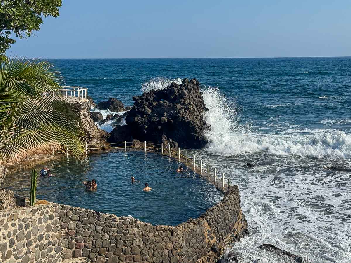View of ocean pool at Atami Escape Resort with a few people swimming and waves crashing in.